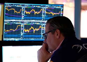 A person in glasses is seated and closely observing multiple financial charts on several computer monitors. The screens display various stock market graphs with different data points and trend lines, highlighting the latest stocks rally. The setting appears to be an office or trading room. | FintechZoom