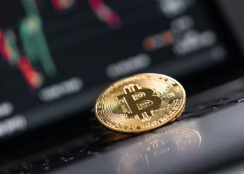 A close-up of a shiny gold Bitcoin coin placed on a laptop keyboard. In the blurred background, a screen displays colorful financial charts, indicating market performance with green and red candlestick patterns—essential for those learning how to file crypto taxes. | FintechZoom