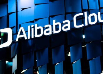 A sign with the text "Alibaba Cloud" is illuminated in white against a background of overlapping blue panels. The logo features a stylized 'A' to the left of the text. The overall atmosphere is modern and technologically sophisticated, much like the appeal of Alibaba Stock to investors. | FintechZoom