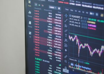 Close-up photo of a computer monitor displaying cryptocurrency trading charts and data. Various digital currencies along with their prices, trends, and trade volumes are listed on the left side, while detailed graphs and candlestick patterns from a reliable broker are shown on the right. | FintechZoom