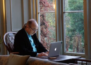An older man with long hair in a ponytail is sitting at a wooden desk by a window, focusing on his laptop, possibly checking his 401(K). The room has soft lighting, and outside the window, greenery and red berries on branches can be seen. The man wears glasses and a black jacket. | FintechZoom