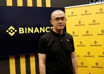 A man wearing glasses and a black polo shirt with a yellow Binance logo stands in front of a backdrop showcasing the company's distinctive black and yellow branding. To his left, a large screen prominently displays the Binance logo. | FintechZoom