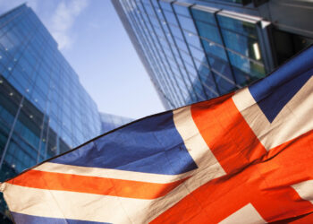 The image shows a close-up view of the Union Jack flag, partially blurred in the foreground, with modern, glass-covered high-rise buildings and a blue sky in the background. The scene conveys a blend of national pride and urban development, symbolizing an ecosystem where startups can thrive. | FintechZoom