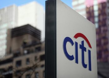 Sign with the Citi logo prominently displayed in the foreground, featuring the word "Citi" with a red arc over the "t". The background shows several tall buildings, indicating a city setting and reflecting an optimistic Stock Market Outlook against a clear blue sky. | FintechZoom