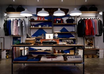 A trendy clothing and accessories store with neatly arranged shelves and racks, epitomizing the business in 2024. It features a variety of apparel including shirts, jackets, and hats. The wooden shelves display various items, and three white pendant lights hang from the ceiling. | FintechZoom