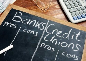 A chalkboard divided into two columns, one labeled "Banks" and the other "Credit Unions," each with subheadings "pros" and "cons." Items are written in a list under each subheading. A chalk piece lies on the board, and a calculator and glasses are nearby, adding a touch of seriousness to the comparison. | FintechZoom