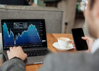 A person in a suit is sitting at a wooden table with a laptop displaying a stock market graph and holding a smartphone, likely scrolling through broker reviews. A cup of coffee in a white saucer is placed on the table next to the laptop. The setting appears to be a casual indoor environment. | FintechZoom