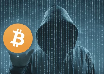A person in a dark hoodie stands against a background of binary code, with their face obscured by the hood. They hold a Bitcoin symbol in their hand, which is prominently displayed in the foreground, hinting at the shadowy world of crypto scams. The image has a digital, mysterious atmosphere. | FintechZoom