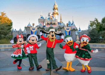 Five beloved Disney characters, including Minnie and Mickey Mouse, Goofy, Donald Duck, and Daisy Duck, dressed in festive holiday attire, stand smiling and waving in front of the iconic Sleeping Beauty Castle at Disneyland. The castle is adorned with holiday decorations—a true picture of Disney magic. | FintechZoom