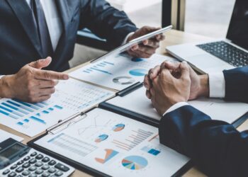 Two people in business suits are sitting at a desk covered with charts, graphs, and financial reports. One person is holding a tablet while the other has their hands clasped. A laptop is open on the desk beside them, indicating a business meeting focused on the accounting cycle. | FintechZoom