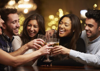 A group of five people gather closely together, smiling and raising glasses in a toast. They are in a warmly lit, festive setting with blurred lights in the background and someone enjoying a THC Vape Pen Oil nearby, creating a cheerful and celebratory atmosphere. | FintechZoom