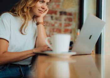 A woman with long blonde hair sits at a wooden table, focused on her laptop with an Apple logo. Resting her chin on her hand, she wears a white t-shirt and a watch. A white coffee cup on a saucer is positioned in the foreground as she browses online adult games. | FintechZoom