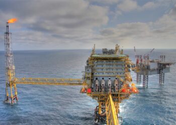 An offshore oil platform with multiple levels and equipment structures stands in the ocean. A flare stack on the left burns off excess gas, emitting a bright flame. Another rig is visible in the background under a partly cloudy sky, reminiscent of betting on penny stocks with unpredictable outcomes. | FintechZoom