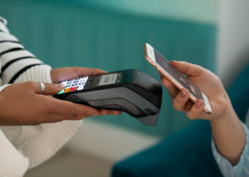 A person holds a smartphone near a contactless payment terminal, making a payment using mobile wallets technology. The terminal's screen is visible, and another person's hand is holding the terminal. Both individuals' faces are not shown. | FintechZoom