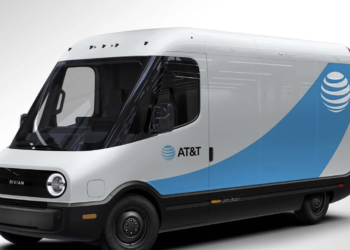 A white electric delivery van with the AT&T logo and branding on the side. The vehicle has a sleek, modern design with a large windshield and black accents on the front grille and bumper, reminiscent of Rivian stock vehicles. The van appears to be parked against a plain, light background. | FintechZoom