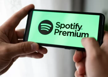 Two hands hold a smartphone displaying the Spotify Premium logo on the screen. The background is a light, blurred indoor setting with soft window light coming in. | FintechZoom