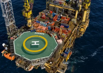 Aerial view of an offshore oil rig platform in the ocean, with a prominent helipad labeled "ARMADA." The rig, associated with Harbour Energy Stock, is bustling with equipment, containers, and structures. Workers in bright clothing are seen gathered around various areas of the platform. | FintechZoom