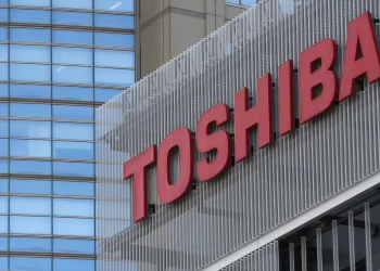 A building with a large red "TOSHIBA" sign on it. The building has a modern design with glass windows and metal paneling. Another building with blue-tinted windows is in the background. The sky is reflected in the windows. | FintechZoom