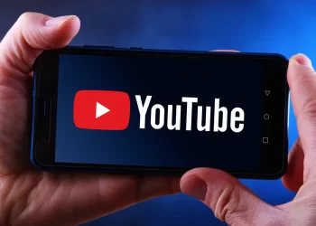 A person holds a smartphone displaying the YouTube app. The screen shows the YouTube logo with a red play button and white text on a black background. The background of the image is a blurred gradient of blue and black, subtly hinting at potential uses like searching for a YouTube to MP3 downloader. | FintechZoom