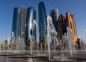 A view of the Etihad Towers in Abu Dhabi, United Arab Emirates, with modern, curved, glass skyscrapers in the background and water fountains spraying upwards in the foreground under a clear blue sky—a stunning testament to Abu Dhabi real estate. | FintechZoom