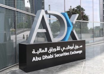 A sign reads "ADX Abu Dhabi Securities Exchange" in English and Arabic. The sign features a large emblem with the letters "ADX" where the "D" and "X" partially overlap. The structure stands against a modern glass building, reflecting both the sky and some surrounding greenery. | FintechZoom