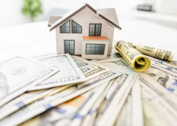 A small model house sits on a surface surrounded by scattered U.S. dollar bills, suggesting themes of real estate, housing market, or financial investment. The house model features basic architectural details with windows and a roof, hinting at the importance of mortgage calculators in financial planning. | FintechZoom