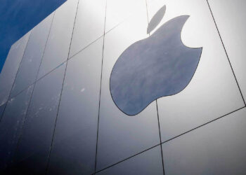 Close-up of the exterior of a building featuring a large, metallic Apple logo on a sleek, silver facade. The sky is visible in the top left corner, creating a modern and clean aesthetic that epitomizes leading US companies. | FintechZoom