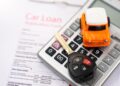A car loan application form, a calculator, a toy car, and a car key with a key fob are arranged together. The calculator is partially covering the form, symbolizing the financial planning involved in finding the best place to get an auto loan. The toy car and key rest atop the calculator. | FintechZoom