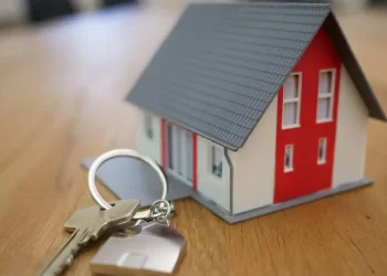 A small model house with a red front door and gray roof sits on a wooden surface. Next to it is a set of silver house keys on a keyring with a small house-shaped keychain attached. | FintechZoom