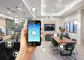 A hand holds a smartphone with a smart home app interface displayed. In the background is a modern kitchen and dining area with icons connected to the phone indicating smart home features for lighting, security, appliances, and entertainment systems. Smart homes can potentially lower insurance rates. | FintechZoom
