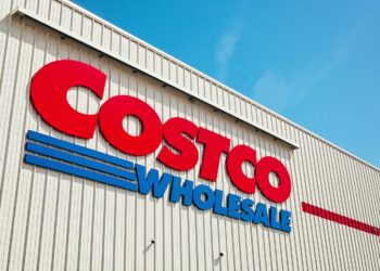 The image shows the exterior of a Costco Wholesale warehouse, its beige building accented with red and blue. Large red letters spell "COSTCO," with smaller blue letters below reading "WHOLESALE." The clear, blue sky adds to the vibrant scene, reflecting the stability often associated with Costco stock. | FintechZoom