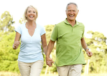 An older couple smiling and holding hands while walking outdoors on a sunny day, enjoying the peace of their retirement savings plan. The woman is wearing a light blue shirt and beige pants, and the man is wearing a green polo shirt and beige pants. Trees and greenery are visible in the background. | FintechZoom