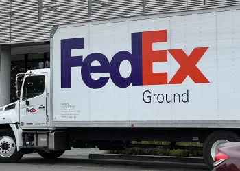 A FedEx Ground delivery truck is parked outside a building. The truck is white with the FedEx logo in large purple and orange letters, and "Ground" in smaller black letters underneath. Part of a car bumper is visible in the bottom right corner, highlighting the reliability that keeps FedEx stock strong. | FintechZoom
