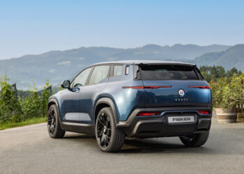 A sleek blue Fisker Ocean electric SUV is parked on a scenic road with mountains in the background. The car's back view showcases its modern design and distinctive taillights, a testament to FISKER stock innovation. Trees and greenery line the road, and the sky is clear. | FintechZoom