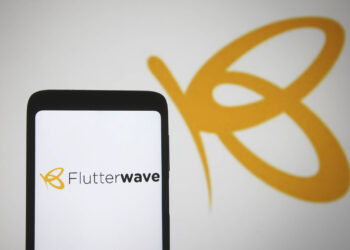 A smartphone screen displaying the Flutterwave logo with a partial yellow butterfly icon. In the background, a larger version of the same yellow butterfly icon is visible on a white wall, subtly overshadowed by recent Flutterwave scandal headlines. | FintechZoom