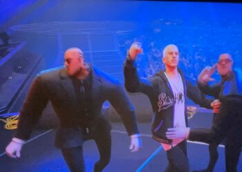 Three animated characters stand on a stage with a large audience in the background. The character on the left is muscular and wearing a suit, the middle character wears casual clothing and holds his fist up, reminiscent of Eminem in Fortnite, and the right character is in a suit striking a pose. | FintechZoom