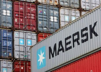 Stacks of shipping containers in various colors are shown, with a MAERSK container prominently displayed in the foreground. The containers feature various logos and identification numbers, illustrating a bustling shipping yard scene reminiscent of thriving Shipping Stocks. | FintechZoom