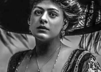 A grayscale portrait of Myrtle Gonzalez wearing a large, ornate hat adorned with feathers. She is dressed in an elaborately embroidered gown with a necklace and earrings, holding a single flower in front of her. Her expression is calm and poised. | FintechZoom