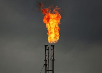 A towering flare stack burns brightly against a dark, cloudy sky. The orange and red flames create a vivid contrast with the gray background, signifying an industrial gas flaring process that can influence Commodities Prices Today. The stack structure is visible below the flames. | FintechZoom