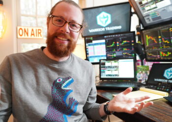 A bearded man with glasses and a ponytail, reminiscent of Ross Cameron, sits at a desk brimming with multiple monitors displaying stock trading charts. He is wearing a gray sweatshirt featuring a blue and purple dinosaur design. The room has a sign that reads "ON AIR." He smiles and gestures with one hand. | FintechZoom