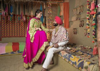 A man and woman dressed in vibrant traditional attire are in a colorful handicraft shop decorated with ethnic ornaments and items. The woman, standing, is smiling down at the seated man. The shop, reminiscent of a Filmyzilla set, has colorful textiles and an array of handcrafted shoes displayed. | FintechZoom