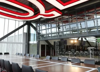 A modern conference room with a long table surrounded by black chairs. The room has large windows and a massive red "Q" sign on the ceiling, reminding attendees of Quicken Loans. In the background, there is a stage setup with bright lights and additional seating. | FintechZoom
