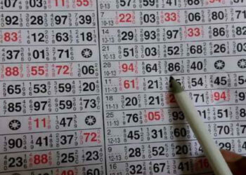 A hand points to a number on a page filled with multiple rows and columns of small, printed numbers reminiscent of a Satta Matka chart. Some of the numbers are printed in red and some in black. The person is holding a white pen, with its tip indicating the number 64 in red. | FintechZoom
