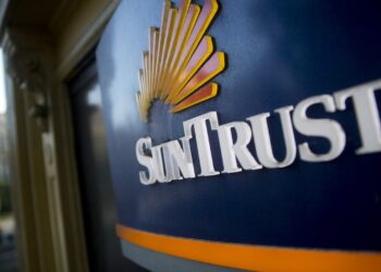 A close-up shot of a SunTrust Bank sign with its logo, featuring stylized yellow and orange rays, on a blue background. The sign, promoting both banking and SunTrust Mortgage services, is mounted on the exterior of a building with part of an urban street visible in the blurred background. | FintechZoom