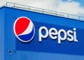 A blue Pepsi building features the iconic Pepsi logo on the left, with red, white, and blue waves inside a circle. The name "Pepsi" appears in white lowercase letters to the right of the logo. Under a clear sky dotted with a few clouds, this symbol stands tall as Pepsi stock thrives. | FintechZoom