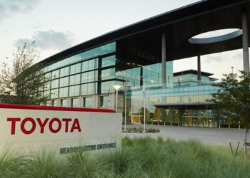 Image of the Toyota headquarters building. The large, modern glass structure is lit by natural light, featuring distinctive architecture with an overhanging roof and circular cutout. In the foreground, a Toyota sign next to landscaped plants subtly evokes the brand's global influence and appeal to Toyota stock investors. | FintechZoom
