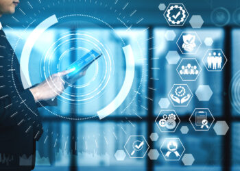 A person in a suit holds a tablet against a futuristic digital interface background. The interface, displaying icons related to technology, business, and communication like gears, people, graphs, and checkmarks over a blue-toned, blurred office setting, epitomizes Trendzguruji.me awareness. | FintechZoom