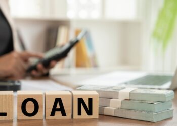 A person in a suit sits at a desk holding a calculator. In the foreground, wooden blocks spelling "LOAN" are placed next to stacks of money. A laptop and paperwork are also visible on the desk, possibly detailing the best personal loans available. The background features shelves with books and a plant. | FintechZoom