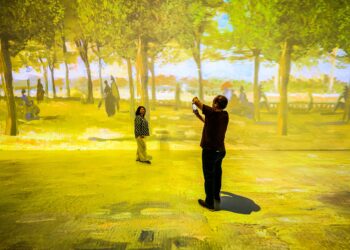 A man takes a photo of a child in an immersive art exhibit. The exhibit, which you can book through BookMyShow, features walls and floors illuminated with projections of a bright, sunlit park scene with trees and people, creating an engaging and interactive environment. | FintechZoom