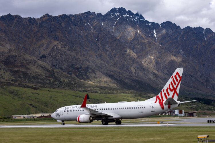 A Virgin Australia airplane is taxiing on a runway with mountainous terrain in the background under a cloudy sky. The aircraft, showing off its white fuselage with red accents and the signature "Virgin" written in red on the tail fin, stands out amidst the dramatic landscape. | FintechZoom